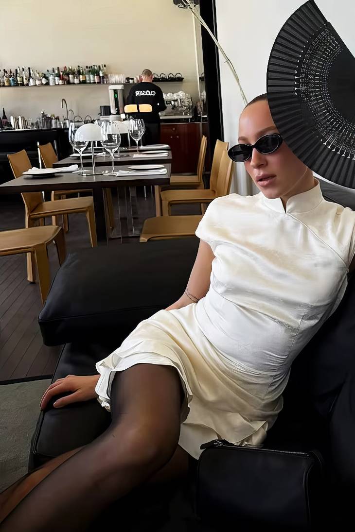 Mujer sentada con qipao mini blanco de jacquard con volantes, cuello alto y mangas cortas, usando gafas de sol y abanico negro en restaurante
